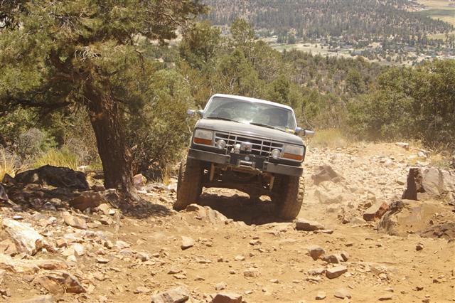 1987 Ford Bronco driving on a steep, rocky off-road trail with trees and a valley in the background