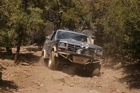 1986 Ford Bronco - A dark blue Ford Bronco navigates a rocky, off-road trail surrounded by trees.