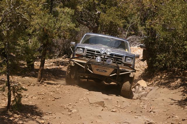 1986 Ford Bronco navigating a steep, rocky off-road trail with trees on either side
