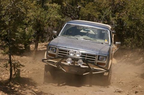 1986 Ford Bronco - This blue SUV, possibly a Ford Bronco, navigates a dusty off-road trail with a custom brush guard and multiple lights.