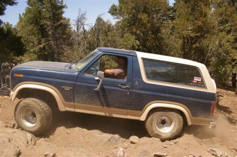 1986 Ford Bronco - A blue Ford Bronco navigates a rough, off-road trail, kicking up dust.