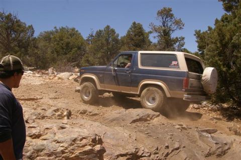 1986 Ford Bronco - A blue and tan Ford Bronco navigates a challenging rocky terrain on a sunny day.