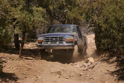 1986 Ford Bronco - A blue 1992 Ford F-150 navigates a challenging, rocky off-road path surrounded by trees.