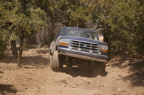 1986 Ford Bronco - A blue Ford Bronco navigates a rocky, dirt trail surrounded by trees.