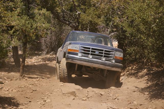 Blue Ford Bronco driving off-road, front 3/4 view. Grille and headlights visible