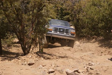 1986 Ford Bronco - A blue Ford Bronco drives through a rocky, wooded off-road path.