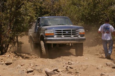 1986 Ford Bronco - A blue Ford F-Series truck navigates a rocky, dusty trail with a person walking alongside.