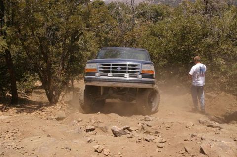 1986 Ford Bronco - A blue Ford Bronco navigates a challenging off-road path, kicking up dust.