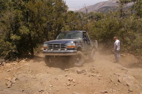 1986 Ford Bronco - A dark blue Ford pickup truck navigates a rocky, dusty trail with a person standing nearby.