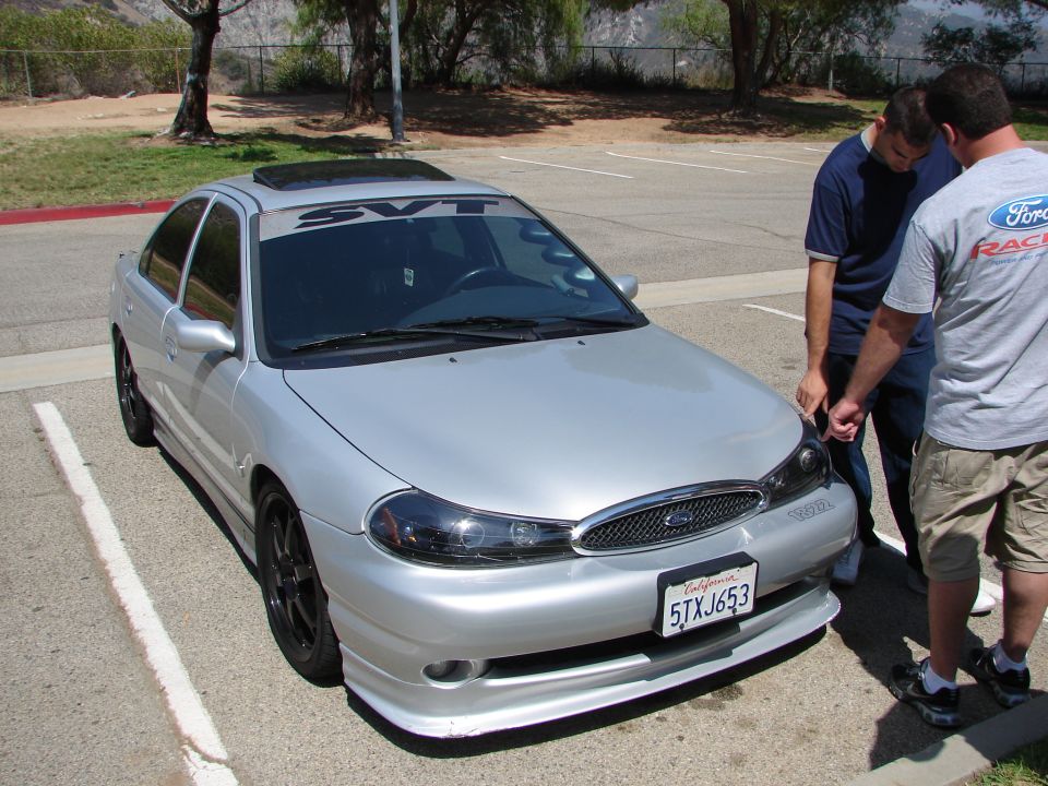 Silver sedan with SVT banner on windshield and aftermarket wheels. Two people are looking at the front of the car