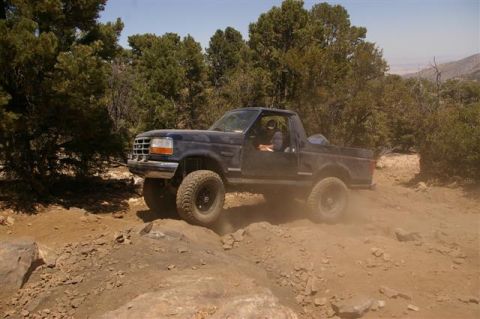 1986 Ford Bronco - A dark blue SUV with lifted suspension and off-road tires navigates a rocky, dusty trail in a wooded, mountainous area.