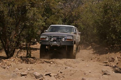 1986 Ford Bronco - A dark-colored Ford Bronco navigates a challenging off-road path, kicking up dust.