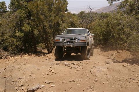 1986 Ford Bronco - A lifted Ford Bronco navigates a challenging dirt path in a wooded, mountainous area.