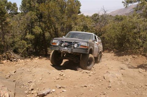 1986 Ford Bronco - A modified Ford Bronco navigates a rocky, off-road path surrounded by trees.