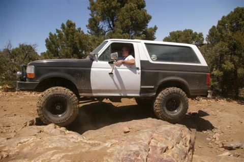 1986 Ford Bronco - A man drives a two-tone black and white Ford Bronco over a rocky obstacle on a dirt trail.