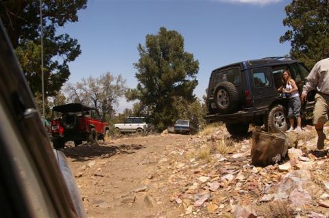 1986 Ford Bronco - Multiple off-road vehicles are seen traversing a , rocky trail surrounded by trees on a sunny day.