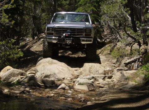 1986 Ford Bronco - A silver 1986 Ford Bronco tackles a challenging, rocky path through a wooded area.