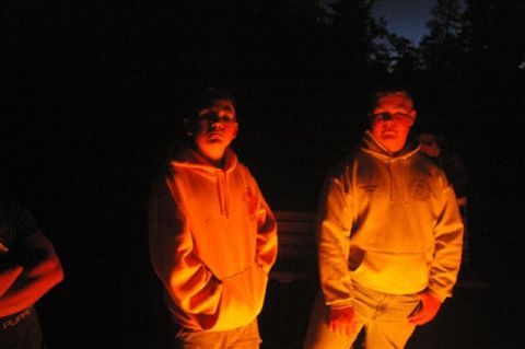 1986 Ford Bronco - Two individuals stand near a campfire, their faces and clothing lit by the flames in the darkness.