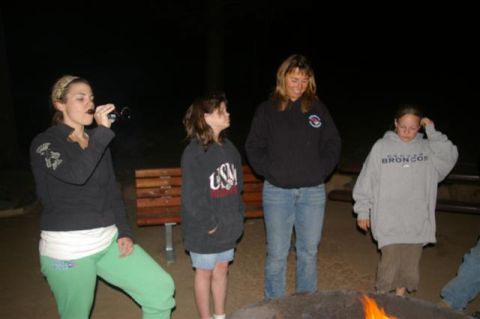 1986 Ford Bronco - Four people stand around a campfire at night, with trees in the background.