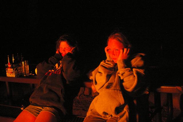 Two people sit by a fire at night, holding their hands to their faces. Bottles of Bud Light are on a table to the left