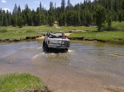 1986 Ford Bronco - White 1986 Ford Bronco convertible navigates a shallow stream surrounded by trees.