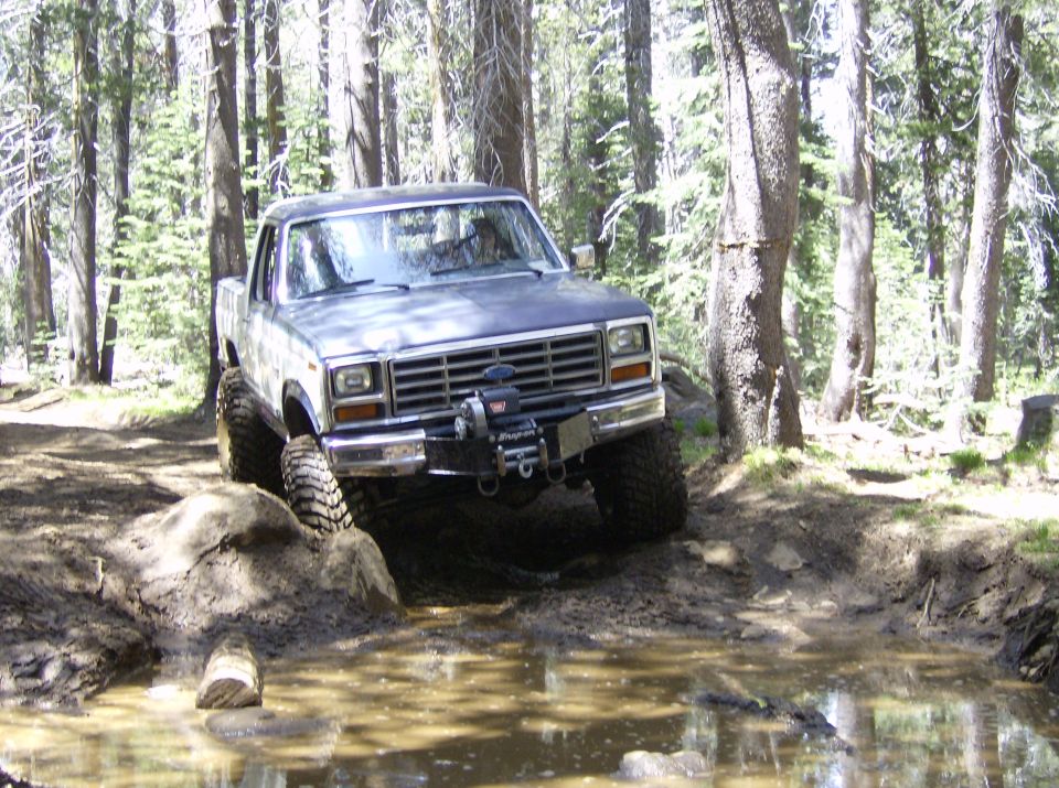 Ford F-Series truck going through mud and water on a wooded trail. Has a winch on the front