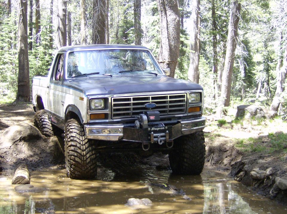 Front view of a modified 1980s Ford pickup truck with large tires and a winch, driving through mud