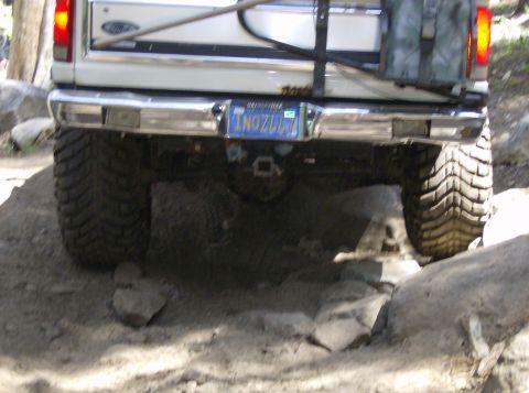 1986 Ford Bronco - A white pickup truck navigates a rocky path, showing its rear end and large tires.