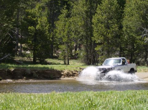 1986 Ford Bronco - A two-tone Ford truck splashes through a shallow stream surrounded by trees and grass.