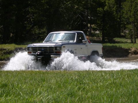 1986 Ford Bronco - A white and blue Ford Bronco navigates through a water crossing, kicking up a large spray.