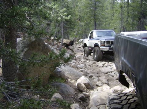 1986 Ford Bronco - Off-road vehicles traverse a challenging rocky path through a wooded area.