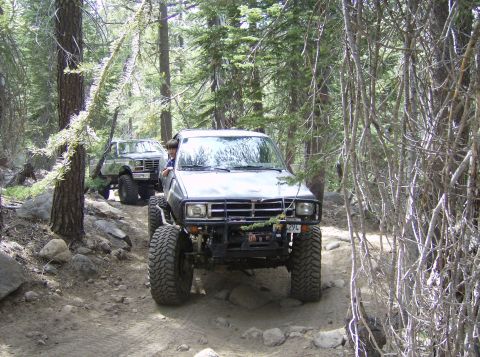 1986 Ford Bronco - Two modified off-road vehicles navigate a narrow, rocky path through a dense forest.