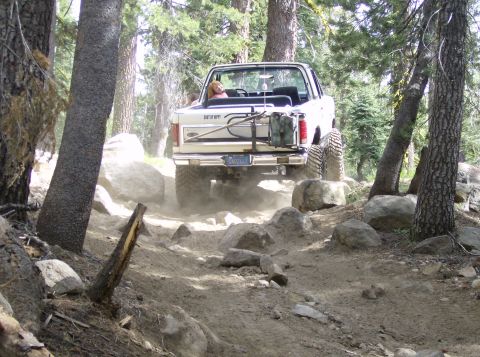 1986 Ford Bronco - A white 1986 Ford Bronco tackles a challenging, rocky trail surrounded by trees.