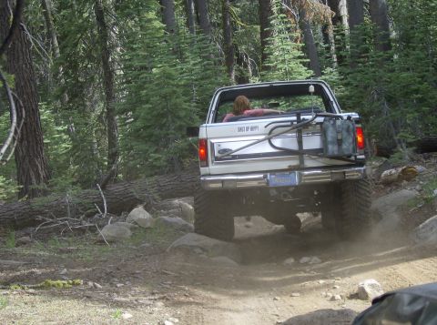 1986 Ford Bronco - White Ford Bronco navigates a challenging off-road trail through a wooded area.