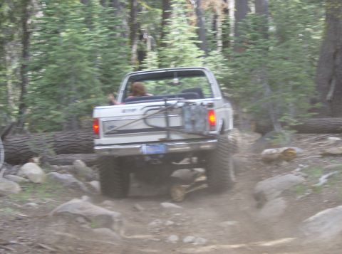 1986 Ford Bronco - A 1986 Ford Bronco navigates a challenging off-road path through a forest with large rocks and fallen trees.
