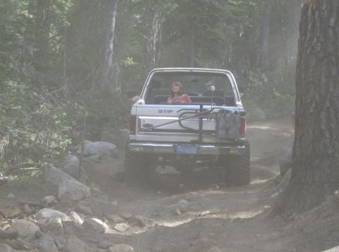 1986 Ford Bronco - A woman rides in the back of a white SUV as it navigates a rocky, dusty trail through a forest.