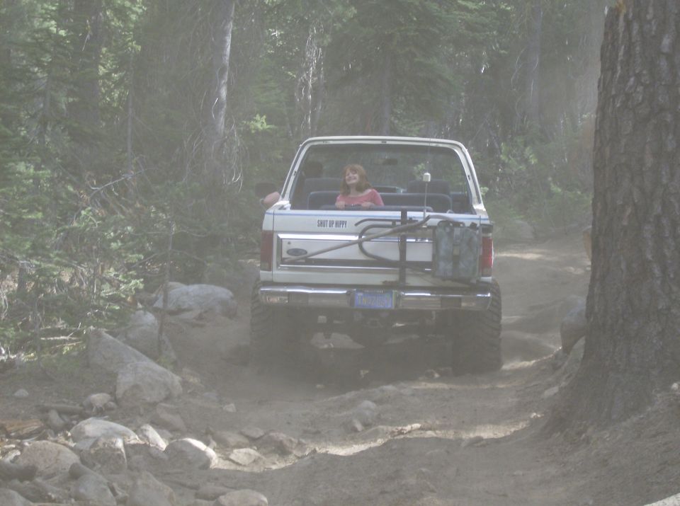 White SUV with a woman in the back, driving on a dusty dirt road through trees. It has a spare tire carrier and a gas can