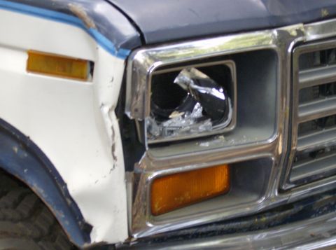 1986 Ford Bronco - Close-up of a damaged front headlight and grille on a white and blue vehicle, showing signs of impact. The grille design suggests an early…