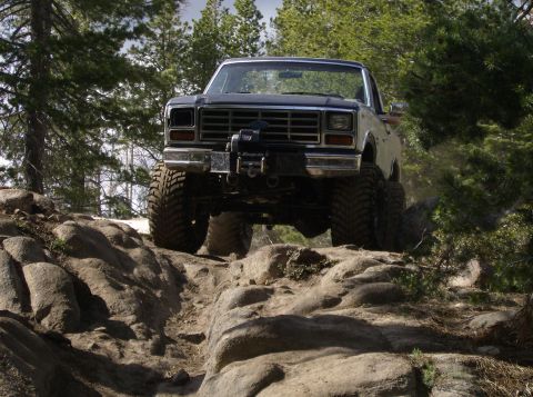 1986 Ford Bronco - This lifted pickup truck, likely from the early 1980s, tackles a challenging off-road course with large tires and a front-mounted winch.
