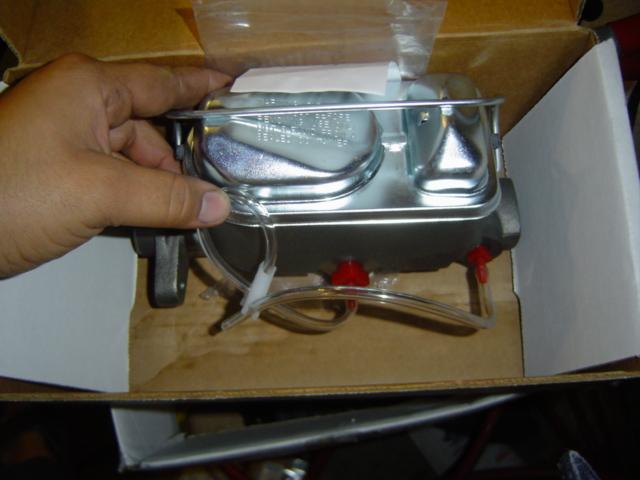 Hand holding a new master cylinder in its box. It has two reservoirs and red caps on the ports