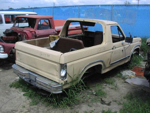 1981 Ford Bronco - A tan 1980 Ford Bronco sits in a junkyard, missing its wheels and doors, with other vehicles in the background.
