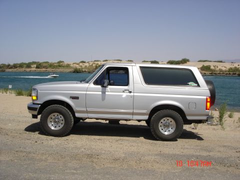 1996 Ford Bronco - This SUV is parked on a sandy area next to a body of water under a clear blue sky.