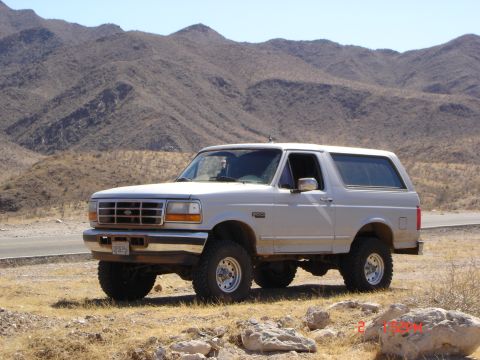 1996 Ford Bronco - This 1996 Ford Bronco is captured in a three-quarter front view, parked on a dirt road with a mountainous backdrop.