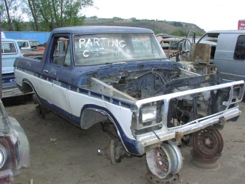 1981 Ford Bronco - A dismantled blue and white Ford truck sits in a junkyard, stripped of its engine and hood.