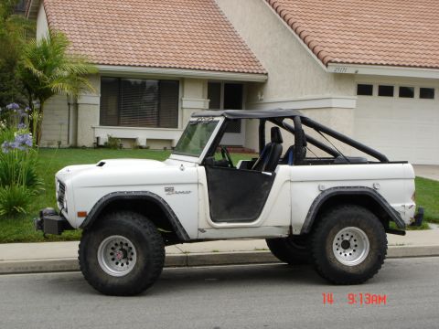 1996 Ford Bronco - White 1970s Ford Bronco with a black soft top and roll cage parked on a street.
