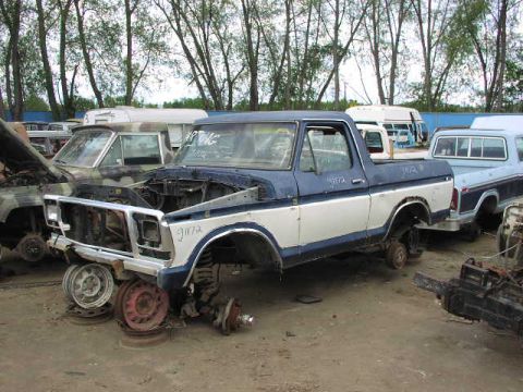 1981 Ford Bronco - Junkyard scene features a two-tone blue and white pickup truck, likely a 1979 model, stripped of its wheels and with its hood open.