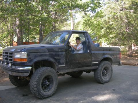 1994 Ford Bronco - A woman drives a dark blue Ford Bronco with a lifted suspension and off-road tires.