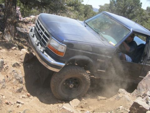 1994 Ford Bronco - A blue 1994 Ford Bronco tackles a challenging off-road trail, kicking up dust as it maneuvers over rocks.