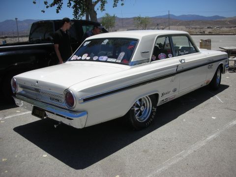 1992 Ford Crown Victoria - A white 1964 Ford Fairlane sedan, modified with racing decals and chrome wheels, is parked outdoors next to a black truck.