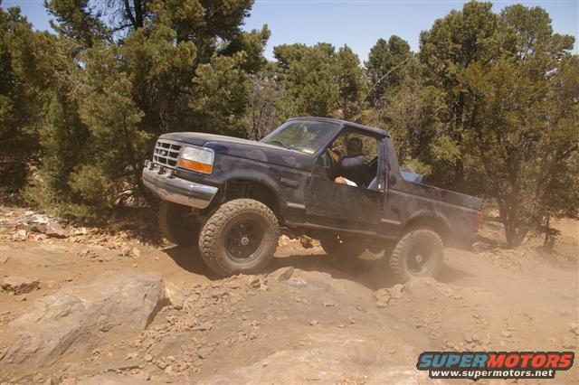 Dark blue Ford Bronco driving over rocks on a dirt trail. Driver visible